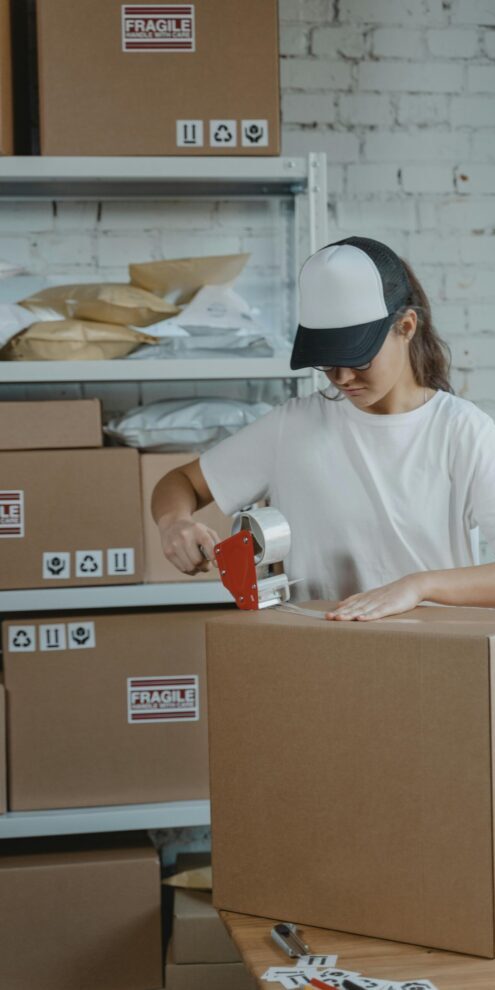 Young female worker sealing cardboard boxes in an indoor warehouse setting.